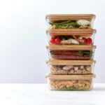 A stack of five glass meal prep containers with wooden lids, each filled with a variety of keto meal prep options like cooked vegetables, grains, and proteins, placed on a white surface.
