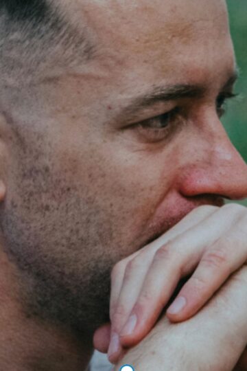 A man with short hair sits outdoors, resting his chin on his hands and looking ahead with a thoughtful expression, as if reflecting on life's challenges.