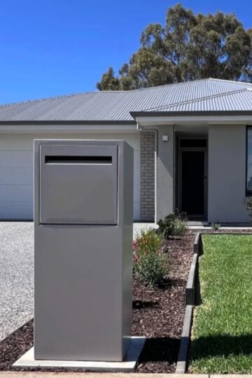 A modern house with a gray metal parcel letterbox in the foreground, a gravel driveway, green lawn, and clear blue sky—highlighting the benefits of secure mail and package deliveries.