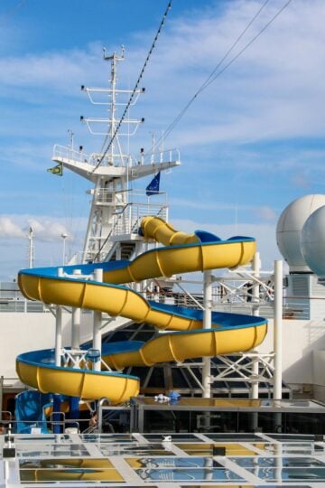 A yellow and blue water slide on the deck of a cruise ship invites fun, with white radar domes and a lounge area in the background—perfect for creating memories on your unforgettable itinerary.