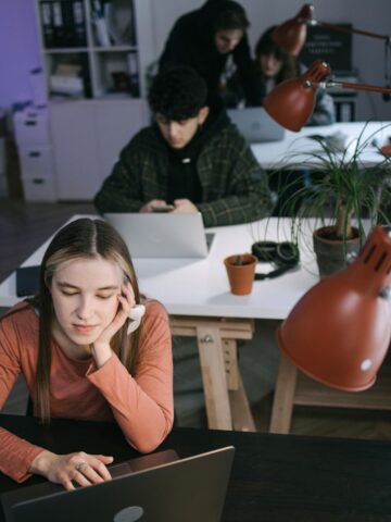A young woman works on a laptop at a desk in the foreground, with three people using laptops at a table in the background in a modern office space.