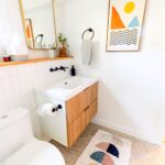 Modern bathroom with a wooden vanity, white sink, toilet, geometric art on the wall, and a textured rug on a light floor-perfect inspiration for your next bathroom renovations.