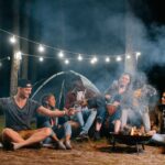 A group of young adults sit around a campfire at night, with string lights overhead and a tent in the background, swapping packing tips and playing instruments as they enjoy the relaxed vibe of car camping.