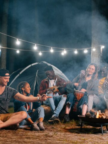 A group of young adults sit around a campfire at night, with string lights overhead and a tent in the background, swapping packing tips and playing instruments as they enjoy the relaxed vibe of car camping.