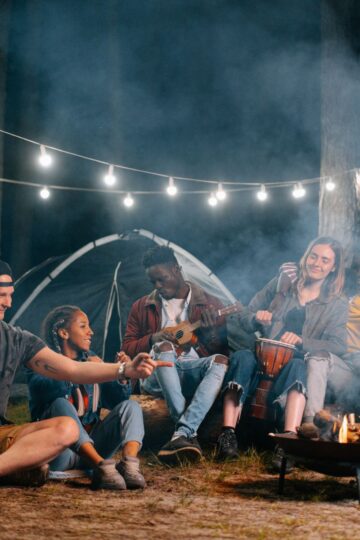 A group of young adults sit around a campfire at night, with string lights overhead and a tent in the background, swapping packing tips and playing instruments as they enjoy the relaxed vibe of car camping.