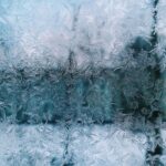 Close-up of intricate frost patterns on a window, resembling frost buildup often seen in a fridge, with a blurred, cool-toned background.
