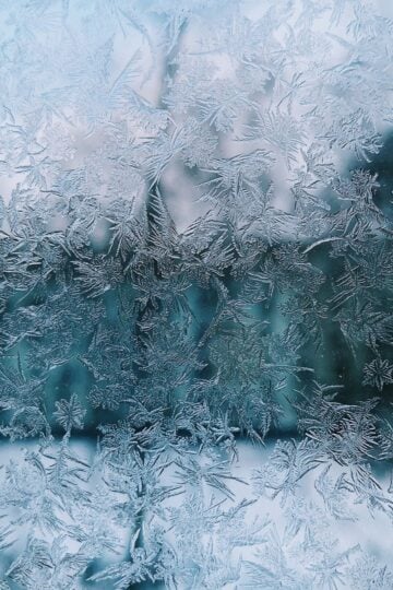 Close-up of intricate frost patterns on a window, resembling frost buildup often seen in a fridge, with a blurred, cool-toned background.