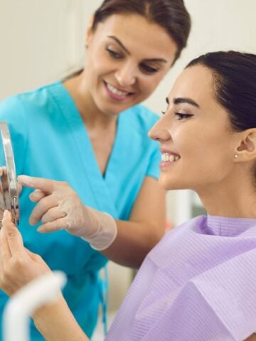 A dentist in blue scrubs shows a female patient her reflection in a handheld mirror during a dental checkup, highlighting how licensed careers in dentistry can be a rewarding college debt alternative.