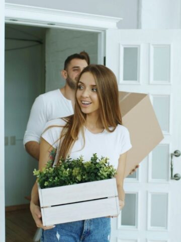 Two people carrying boxes, one with a plant, walk through a doorway, capturing a moment of UK to US relocation or the start of expat life in a new US rental home.