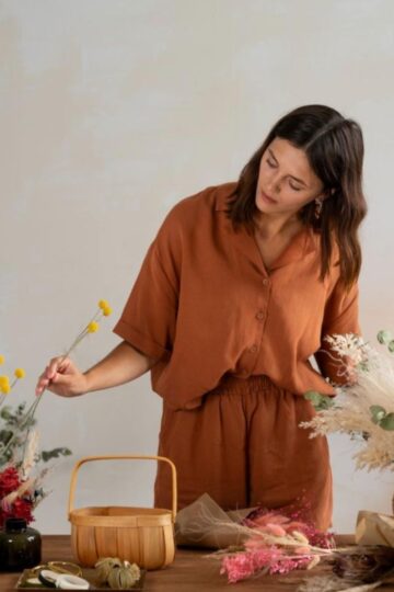 A woman in a brown outfit arranges dried flowers on a table with vases, a wooden basket, and various decorative items, expressing her style and confidence through thoughtful self-care and artistic touches.