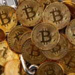 A pile of shiny gold-colored coins with the Bitcoin symbol embossed on them, arranged closely together.