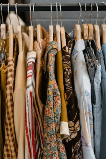 A row of assorted shirts, blouses, and affordable dresses in various colors and patterns hanging on wooden hangers on a clothing rack.