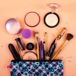 A trending makeup bag with various cosmetics and brushes spilling out, including powders, lipstick, mascara, foundation, and a purple blending sponge, arranged on a peach surface-perfect for what customers want.