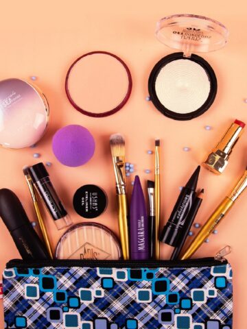A trending makeup bag with various cosmetics and brushes spilling out, including powders, lipstick, mascara, foundation, and a purple blending sponge, arranged on a peach surface-perfect for what customers want.