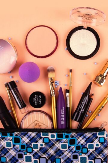 A trending makeup bag with various cosmetics and brushes spilling out, including powders, lipstick, mascara, foundation, and a purple blending sponge, arranged on a peach surface—perfect for what customers want.