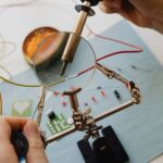 A person uses a soldering iron and magnifying glass to work on an electronic circuit, skillfully addressing hidden wiring issues with wires, resistors, and a circuit board on a blue surface.