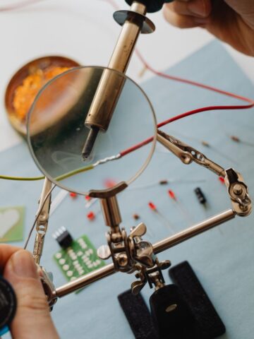 A person uses a soldering iron and magnifying glass to work on an electronic circuit, skillfully addressing hidden wiring issues with wires, resistors, and a circuit board on a blue surface.