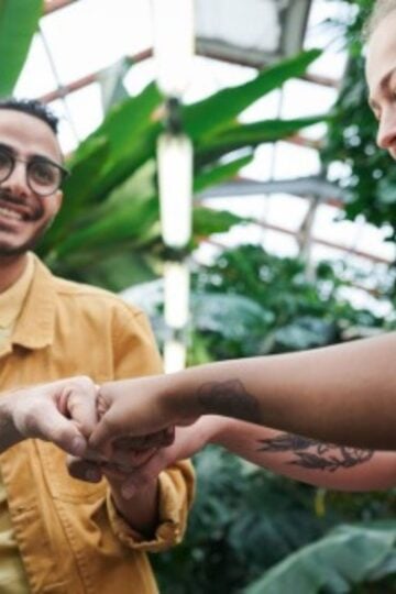 Three people stand in a greenhouse, smiling and joining their hands together in a gesture of teamwork or unity, highlighting the social dimension of health. Large green plants surround them.