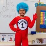 A child wearing a red "Thing 1" costume and blue wig stands in a classroom, smiling and waving. The whiteboard behind says "Hats Off to Reading!" with a Dr. Seuss logo-perfect for Read Across America or fun literacy costume ideas.