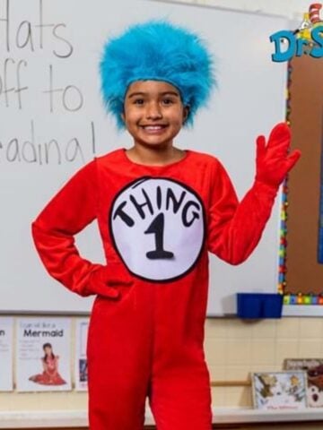 A child wearing a red "Thing 1" costume and blue wig stands in a classroom, smiling and waving. The whiteboard behind says "Hats Off to Reading!" with a Dr. Seuss logo-perfect for Read Across America or fun literacy costume ideas.