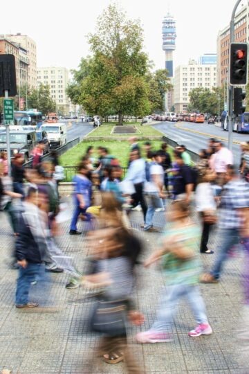 A busy city crosswalk with people from diverse communities walking in both directions, blurred by motion; buildings, trees, and traffic are visible in the background.
