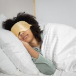 Person wearing a gold sleep mask lies on their side in bed under a white blanket, resting their head on a white pillow-avoiding sleeping wrong to help prevent neck pain.