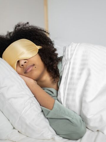 Person wearing a gold sleep mask lies on their side in bed under a white blanket, resting their head on a white pillow-avoiding sleeping wrong to help prevent neck pain.