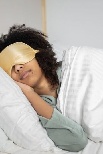 Person wearing a gold sleep mask lies on their side in bed under a white blanket, resting their head on a white pillow—avoiding sleeping wrong to help prevent neck pain.