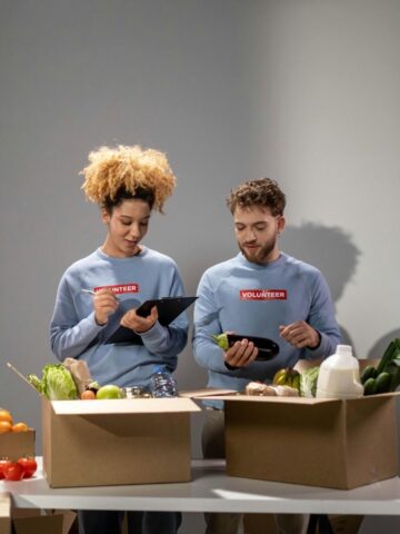 Two people wearing "volunteer" shirts pack fresh produce and groceries into boxes, embodying community development. One holds a tablet while the other lifts an eggplant, working together to build stronger communities.