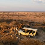 Two people stand beside a safari vehicle on rocky terrain, enjoying a stress-free safari while overlooking a vast savannah landscape under a partly cloudy sky.