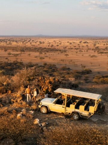Two people stand beside a safari vehicle on rocky terrain, enjoying a stress-free safari while overlooking a vast savannah landscape under a partly cloudy sky.