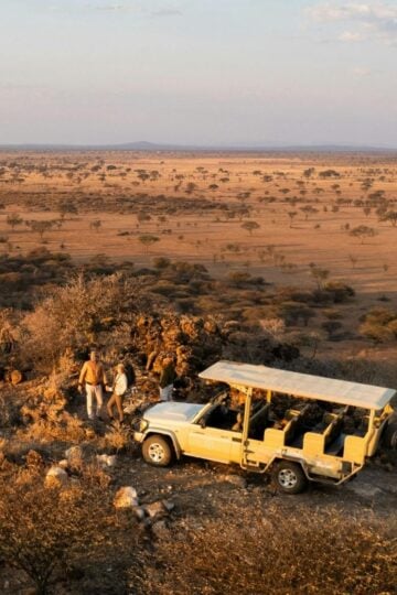 Two people stand beside a safari vehicle on rocky terrain, enjoying a stress-free safari while overlooking a vast savannah landscape under a partly cloudy sky.