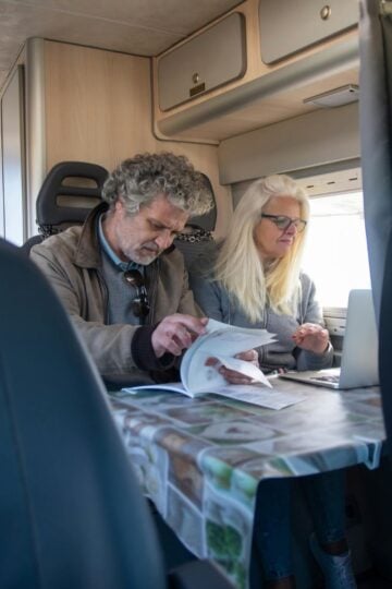 An older man and woman sit at a table in a camper van, looking at papers and a laptop, with natural light coming through the window.