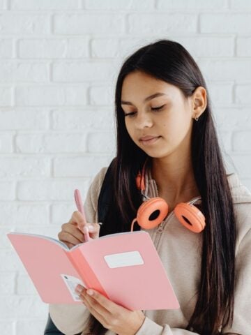 A young woman with long dark hair writes in a pink notebook, immersed in creative writing. She wears a beige hoodie and orange headphones around her neck, seated against a white brick wall.
