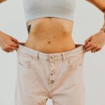 Person wearing a light crop top and holding out the waistband of loose, light-colored pants, revealing a tattoo and showing slim waist-an inspiring look that reflects successful weight loss with possible medical support.