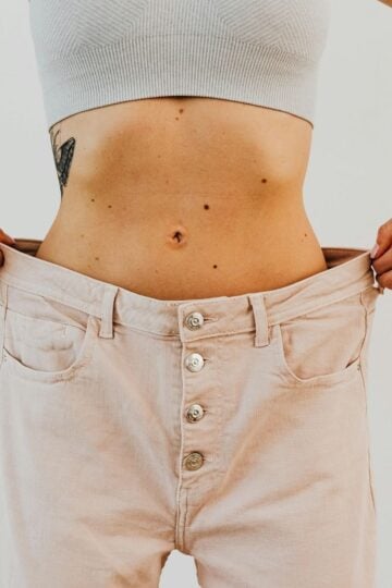 Person wearing a light crop top and holding out the waistband of loose, light-colored pants, revealing a tattoo and showing slim waist—an inspiring look that reflects successful weight loss with possible medical support.