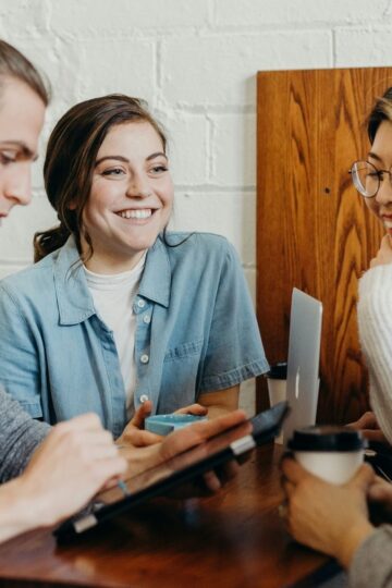 Four lifelong learners sit at a table, talking and smiling, with a tablet, laptop, and coffee cups in front of them—engaged in lively communication.