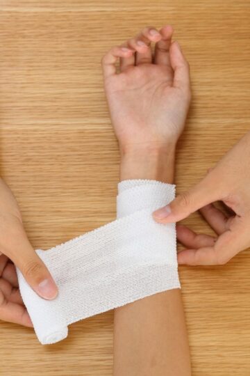 A person wraps a white bandage around another person's wrist on a wooden surface, providing care after a work injury.