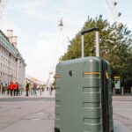 A green hard-shell suitcase stands on a city sidewalk, perfect for city breaks or travel 2026 adventures, with the London Eye and nearby buildings visible in the background.