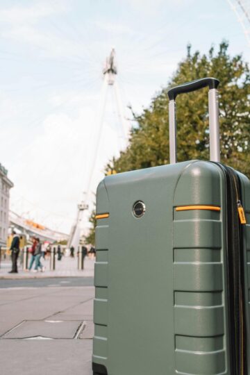 A green hard-shell suitcase stands on a city sidewalk, perfect for city breaks or travel 2026 adventures, with the London Eye and nearby buildings visible in the background.