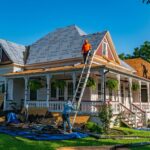 Two workers are re-roofing a home; one stands on a ladder while the other works on the ground. The roof, partially covered with protective material, shows how quickly neglected repair can spark a chain reaction of problems if left unchecked.