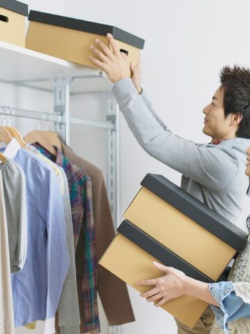 Two people organize a closet by placing brown storage boxes on a high shelf next to hanging clothes.