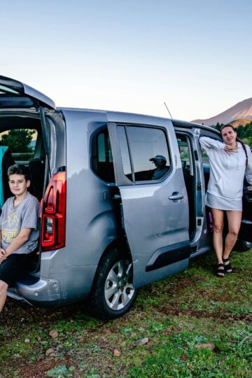 Five people are beside a silver family car with open doors, parked on grass near a mountain in the background, during daylight.