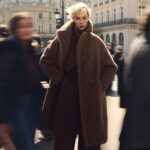A person in a large teddy coat stands still on a city street while blurred figures walk past, with historic buildings in the background.