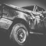 Black and white image of a damaged truck with a dented door and flat tire, wrapped in police tape that reads "POLICE LINE DO NOT CROSS," highlighting the aftermath before truck accident recovery begins.