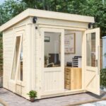 A small, modern wooden garden shed with large windows and an open door sits on a paved patio, designed as a stylish office for working from home with a desk, chair, and storage inside.