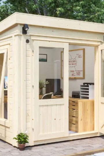 A small, modern wooden garden shed with large windows and an open door sits on a paved patio, designed as a stylish office for working from home with a desk, chair, and storage inside.