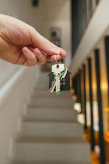 A hand holds a set of keys in front of a staircase inside a modern home, symbolizing a fast home purchase and the excitement of speeding up the home buying process.
