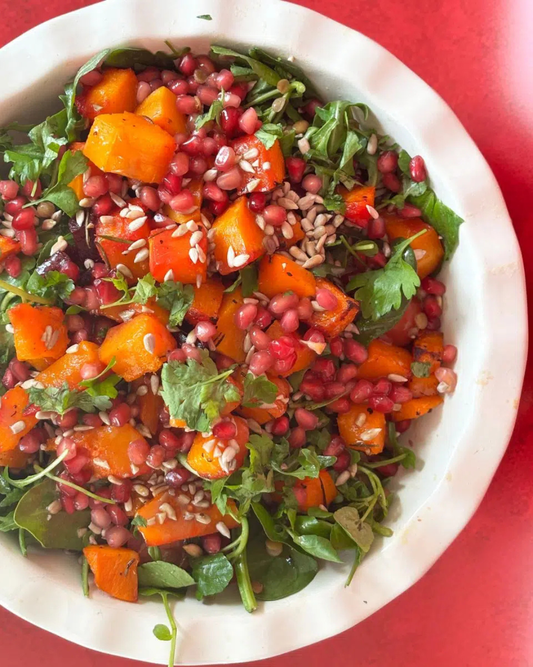 A bowl of salad with roasted orange squash, pomegranate seeds, leafy greens, fresh herbs, and sunflower seeds on a red surface-perfect for Mother's Day lunch ideas or as a fresh addition to your favourite recipes.