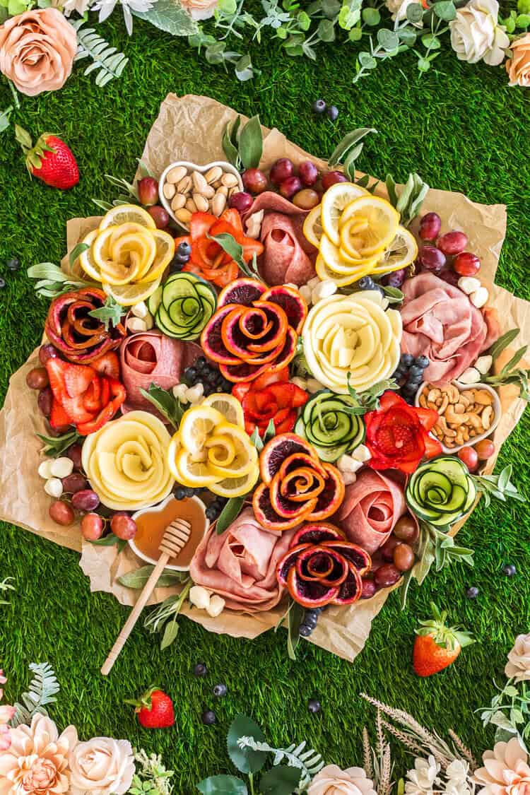 Celebrate With These Recipes for Lunch Ideas on Mother's Day A charcuterie board on greaseproof paper with meats, cheeses, nuts, fruit, and vegetables arranged as flower shapes, surrounded by greenery and flowers on grass.